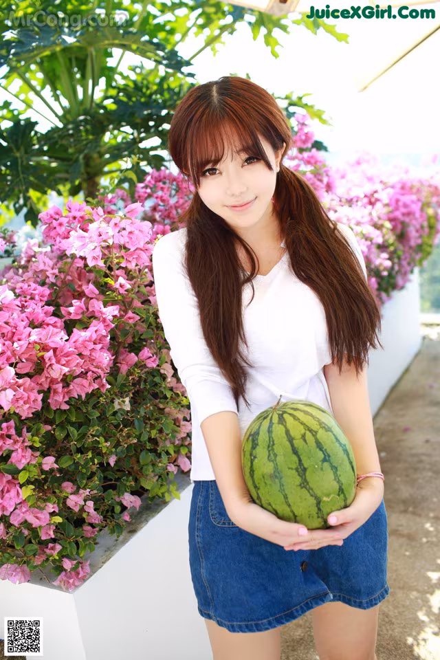 A young woman holding a watermelon in front of flowers.