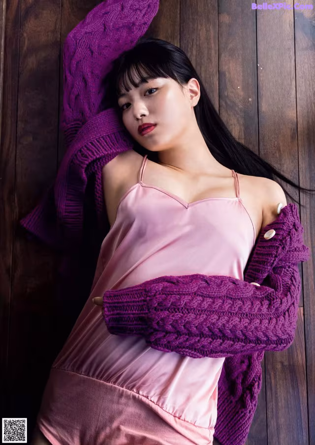 A woman in a pink dress and purple sweater leaning against a wooden wall.
